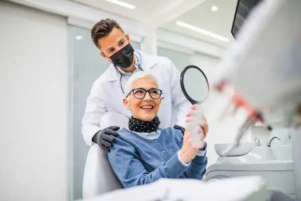 An old woman looking in the mirror and smiling while the dentist stands behind her dental chair.